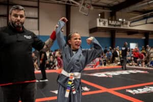 A young girl in a gray BJJ gi celebrates a victory as a NAGA referee raises her arm during a tournament.